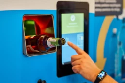 męska dłoń wrzuca szklaną butelkę do butelkomatu powiązanego z polskim systemem kaucyjnym